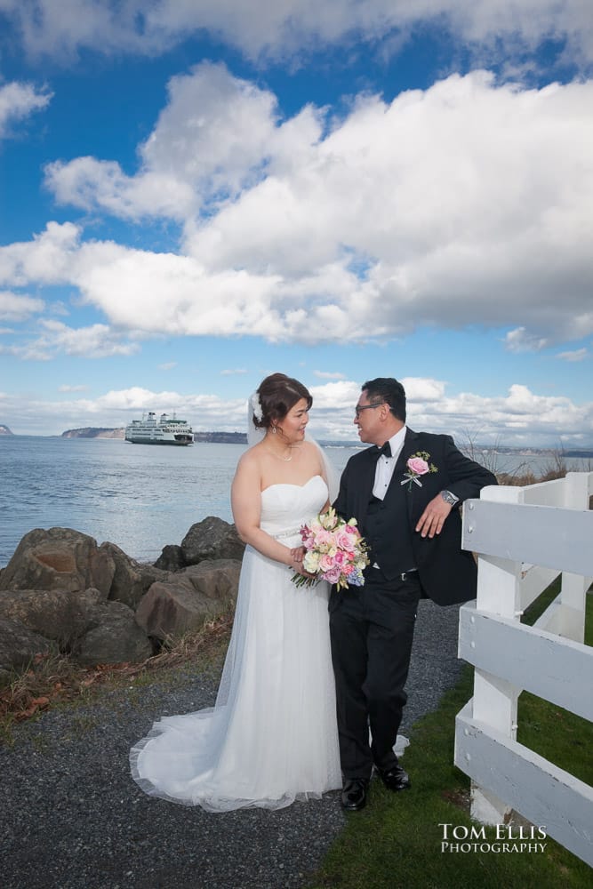 Bride and groom stand together along the edge of the water, with a ferryboat in the background
